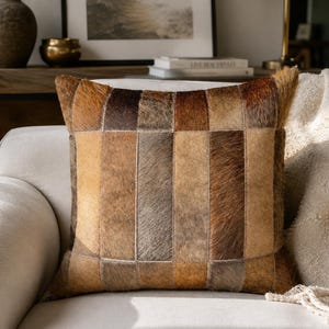 May include: A square throw pillow featuring a patchwork design of vertical rectangular panels made from natural cowhide. The surface displays a varied color palette of light tan, medium brown, dark chocolate, and greyish-brown tones.