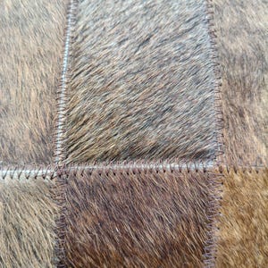 May include: A patchwork of rectangular panels made from natural cowhide with fur texture. The panels feature varying shades of brown, tan, and dark gray, joined together by visible dark brown stitching along the edges of each individual piece.