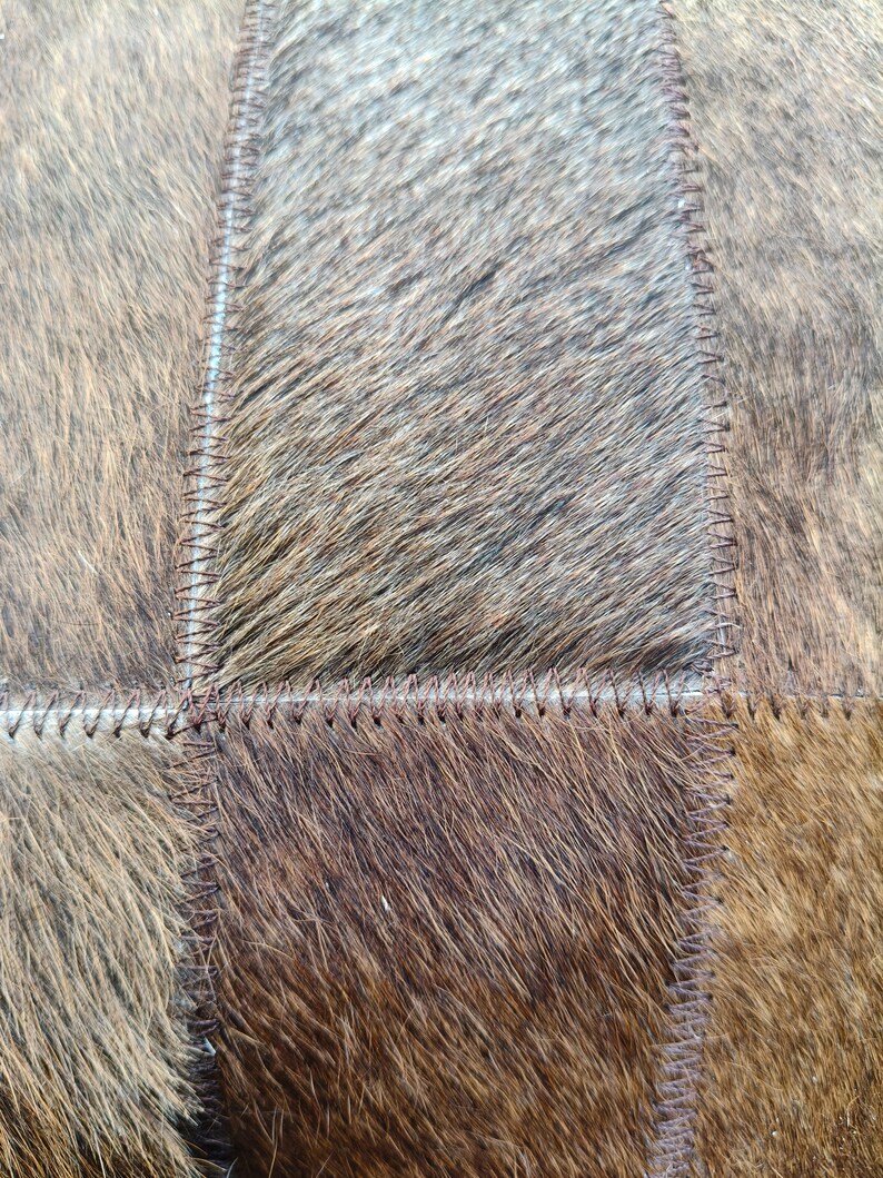 May include: A patchwork of rectangular panels made from natural cowhide with fur texture. The panels feature varying shades of brown, tan, and dark gray, joined together by visible dark brown stitching along the edges of each individual piece.