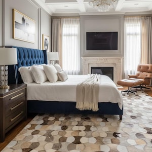 May include: A bedroom featuring a tufted navy blue upholstered bed with white bedding and a beige throw blanket. A patterned cowhide rug with circular shapes in shades of brown, tan, and white covers the floor. A brown leather lounge chair sits nearby.