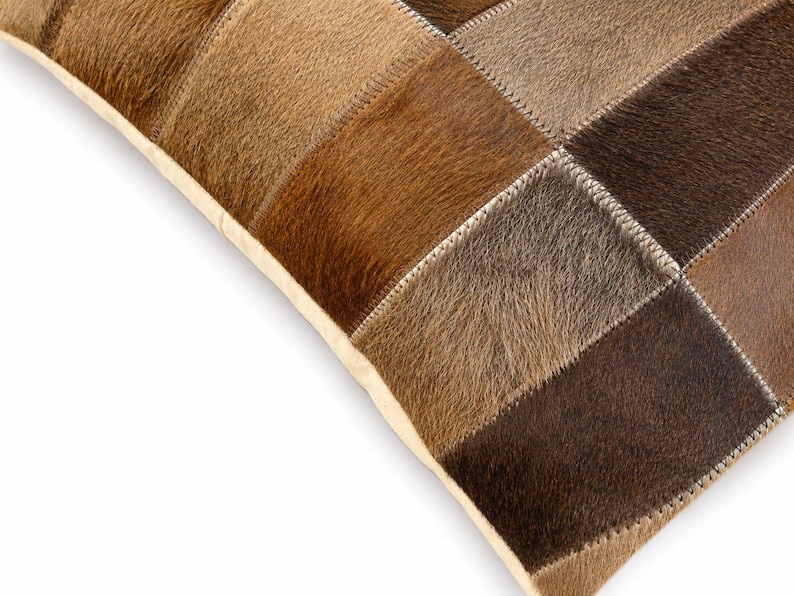 May include: A square throw pillow constructed from a patchwork of rectangular hide panels in varying shades of brown, tan, and dark chocolate. The individual pieces are joined together with visible white stitching along the seams.