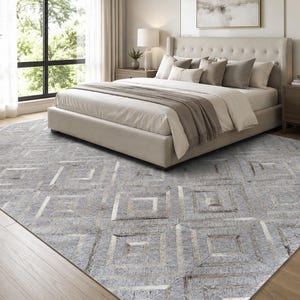 Puede incluir: Una alfombra rectangular grande con un patrón geométrico repetitivo de cuadrados anidados en tonos gris claro, plata y blanco. La alfombra tiene una superficie texturizada con un brillo metálico y está colocada sobre un suelo de madera clara.
