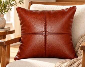 May include: A square throw pillow made of smooth, reddish-brown faux leather. The pillow features a prominent cross-shaped seam design running vertically and horizontally through the center, creating four distinct quadrants on the front surface.