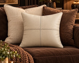May include: A square, cream-colored faux leather throw pillow featuring a central cross-stitch seam detail. The pillow is positioned in front of a textured beige throw pillow and a dark brown throw pillow on a matching dark brown sofa.