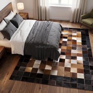 Può includere: Un tappeto rettangolare caratterizzato da un design patchwork di piastrelle quadrate nei toni del marrone, beige, nero e bianco. Il tappeto è posizionato su un pavimento in legno in una camera da letto, con il testo "indianglobalrugs" visibile al centro.