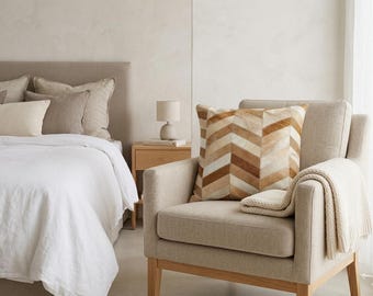May include: A square throw pillow featuring a chevron pattern made of alternating strips of tan, brown, and cream-colored hide. The pillow is positioned on a light beige upholstered armchair with wooden legs and a draped cream-colored knit throw blanket.