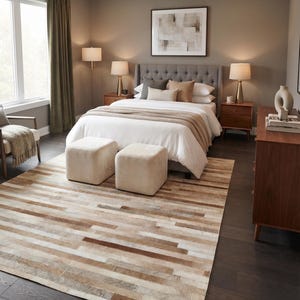 May include: A rectangular area rug featuring a patchwork design of horizontal strips in varying shades of cream, beige, tan, and dark brown. The rug is positioned on a dark wood floor in a bedroom setting, showcasing a textured, multi-tonal pattern.