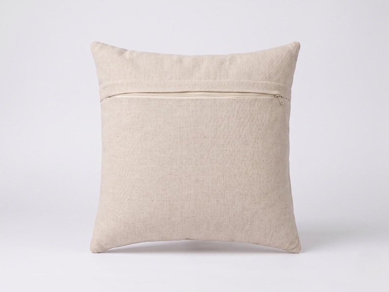 May include: A square throw pillow cover in a solid light beige color. The cover features a horizontal zipper closure positioned across the upper portion of the back side, with a visible metal zipper pull tab on the right side of the fabric.