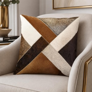 May include: A square throw pillow featuring a geometric patchwork design made of cowhide panels in shades of cream, tan, dark brown, and grey. The panels are arranged in a diagonal pattern with visible stitching along the seams of the hide.