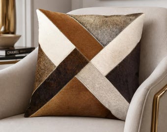 May include: A square throw pillow featuring a geometric patchwork design made of cowhide panels in shades of cream, tan, dark brown, and grey. The panels are arranged in a diagonal pattern with visible stitching along the seams of the hide.