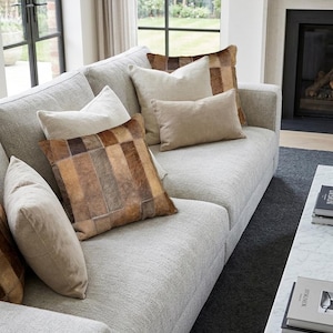 May include: A light gray textured sofa featuring an assortment of throw pillows. The collection includes solid beige square and rectangular cushions alongside square pillows with a brown and tan patchwork hide pattern in various vertical stripes.
