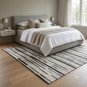 May include: A rectangular area rug featuring a pattern of horizontal stripes in varying shades of grey, white, and black. The rug is positioned on a light-coloured wooden floor beneath a bed with a grey upholstered headboard and white bedding.