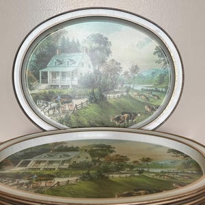 May include: A stack of round metal trays featuring a printed pastoral scene of a white house, a horse-drawn carriage, and grazing cattle in a green landscape. The trays have a metallic silver-toned rim and a smooth, glossy finish on the surface.