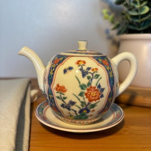May include: A cream-coloured ceramic teapot with a matching saucer, featuring a hand-painted floral pattern in orange, blue, and green. The teapot has a curved spout, a handle, and a lid, decorated with intricate botanical motifs and blue borders.
