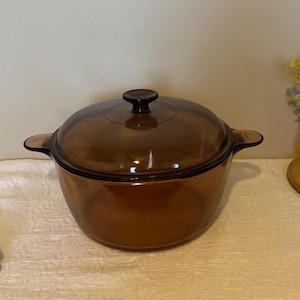 May include: A round, amber-coloured glass cooking pot with a matching transparent glass lid. The pot features two integrated side handles and a central knob on the lid. The glass is translucent, allowing light to pass through the brown-tinted material.
