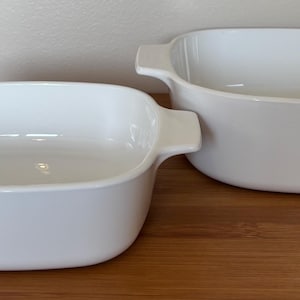 May include: Two white ceramic baking dishes with rounded corners and integrated side handles. The dishes feature a smooth, glossy finish and a deep, open interior design, providing a clean and functional look for kitchen preparation and serving.