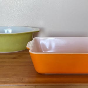 May include: A round, light green ceramic baking dish with side handles and a white interior, paired with a square, bright orange ceramic baking dish featuring side handles and a light pink interior, both designed for oven use and food preparation.