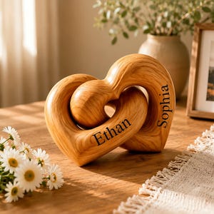 Puede incluir: Una escultura de madera de color marrón claro con dos formas de corazón entrelazadas. El nombre Ethan está grabado en el corazón izquierdo y el nombre Sophia en el derecho. La veta de la madera es visible en toda la superficie lisa y pulida.