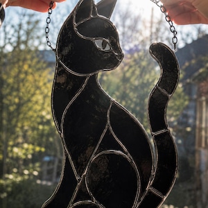 May include: A black stained glass cat suncatcher featuring a sleek silhouette with segmented glass pieces joined by silver metal solder lines. The cat is depicted in a seated position with its tail curved upward, suspended by a thin metal chain.