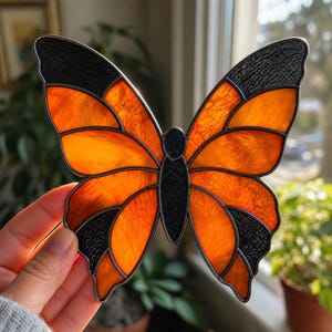 May include: A stained glass butterfly suncatcher featuring vibrant orange translucent wings with textured black glass borders and a central black glass body. The piece is constructed with dark metal soldering lines separating each glass segment.
