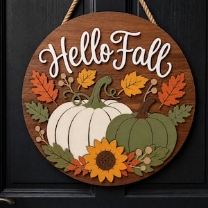 Könnte beinhalten: Ein rundes Wand-Schild aus Holz mit dem Schriftzug Hello Fall in weißer Schreibschrift. Das Design zeigt einen großen weißen Kürbis, einen kleineren salbeigrünen Kürbis, eine gelbe Sonnenblume sowie orangefarbene und grüne Herbstblätter.