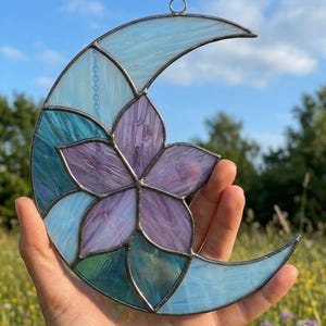 Peut inclure: Un attrape-soleil en forme de croissant de lune en verre teinté, composé d'une fleur centrale violette à cinq pétales entourée de segments de verre bleu clair et sarcelle irisé, assemblés par des lignes de soudure en métal sombre.