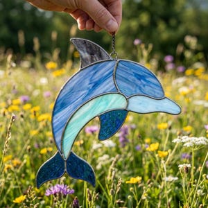 May include: A stained glass dolphin suncatcher featuring various shades of blue, teal, and white glass pieces joined by dark metal solder lines. The dolphin is depicted in a leaping pose with a small metal chain attached to the top fin for hanging.