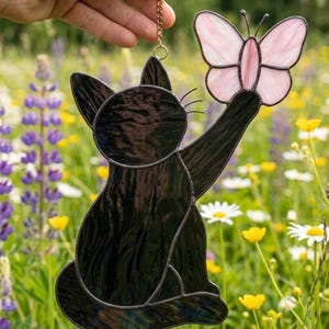 Peut inclure: Un attrape-soleil en vitrail représentant un chat noir tendant la patte vers un papillon rose clair. Le chat et le papillon sont composés de pièces de verre individuelles assemblées par des lignes de soudure en métal sombre, suspendus par une chaîne dorée.