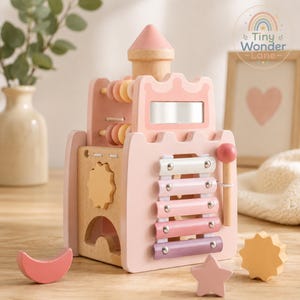 May include: A wooden activity cube shaped like a castle in shades of pink, cream, and natural wood. It features a xylophone with a mallet, a mirror, abacus beads, and a sun-shaped gear. Includes separate wooden moon, star, and sun shapes.