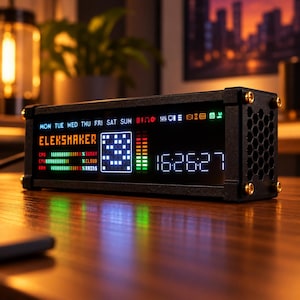 May include: A rectangular black electronic desktop clock with a digital display labelled ELEKSHAKER. The screen shows the days of the week, CPU and ARM usage bars, weather icons, a pixelated graphic, and the current time in white digits.