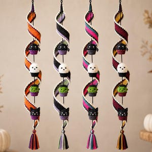May include: Four vertical hanging decorations featuring crocheted spiral ribbons in orange, purple, pink, and burgundy. Each ribbon holds four small crocheted figures: a black spider, a white ghost, a green monster, and a black cat, ending in a tassel.