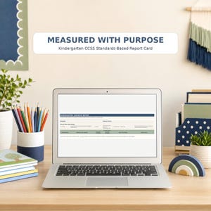 May include: A silver laptop displaying a Kindergarten Learning Report on its screen. Above the laptop, a white rectangular label reads MEASURED WITH PURPOSE Kindergarten CCSS Standards-Based Report Card in dark blue and black text.