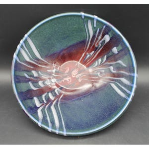 May include: A round ceramic bowl featuring a deep green and blue base glaze with a central reddish-brown circular accent. The surface is decorated with radiating white drip patterns that extend from the centre towards the outer rim.