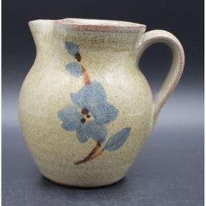 May include: A small ceramic pitcher with a speckled beige glaze and a rounded body. The pitcher features a hand-painted blue flower design with brown stems and leaves on the front. It has a curved handle on the right side and a small pouring spout.