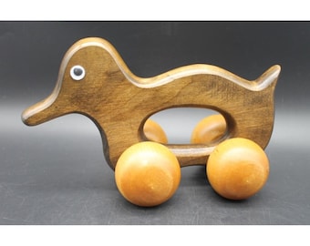 Könnte beinhalten: Ein dunkelbraunes Holzspielzeug in Entenform mit einem glatten, geschwungenen Körper und einer Aussparung in der Mitte. Die Ente hat ein einzelnes weiß-schwarzes Wackelauge am Kopf und ist auf vier hellen, runden Holzrädern montiert.