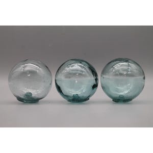 May include: Three spherical glass floats arranged in a horizontal row. Each orb features a translucent, pale blue-green tint, a smooth yet slightly irregular surface texture, and a small, raised glass knob at the base of each sphere.