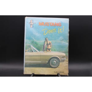 May include: A hardcover book titled Mustang Does It! by Ray Miller. The cover features a tan convertible car, a person in a black bikini standing by the vehicle, and a small Mustang logo in the upper left corner against a light blue background.