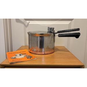 May include: A stainless steel Revere Ware pressure cooker with a copper-clad base, two black plastic handles, and a metal pressure regulator on the lid. An orange instruction manual titled Revere Ware Pressure Cooker is placed beside it.