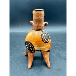 Op de afbeelding: Een terracotta vaas met een hoge hals, twee kleine gekrulde handvatten en drie poten. Het lichaam heeft een warme oranje en bruine afwerking met twee opvallende zwarte cirkelvormige vlakken met witte driehoekige patronen aan de zijkanten.
