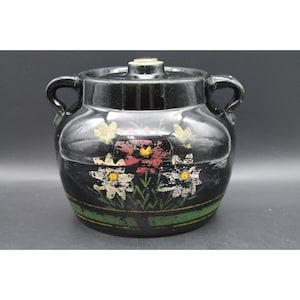 Op de afbeelding: Een zwarte keramieken pot met twee zijhandvatten en een bijpassend deksel. De buitenkant is voorzien van een handgeschilderd bloemmotief met witte, rode en gele bloemen, geaccentueerd door een horizontale groene en gele band nabij de basis.