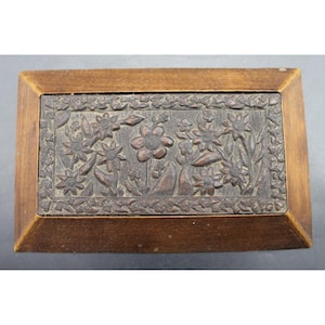 May include: A rectangular wooden box featuring a carved lid with a central floral motif surrounded by a detailed border of small leaves and flowers. The box has a smooth, angled frame and a dark brown finish with visible wood grain texture.