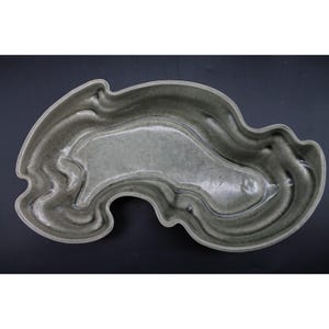 May include: An irregular, organic-shaped ceramic dish with a glossy, muted sage green glaze. The surface features deep, flowing indentations and raised ridges that create a rippled, pond-like appearance with subtle variations in color depth.