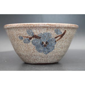 May include: A speckled ceramic bowl with a light beige base and brown flecks throughout. The exterior features a hand-painted design of a blue flower with dark brown stems and leaves. The bowl has a wide, open top and a tapered, rounded base.