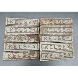 May include: Ten rectangular metal printing blocks arranged in two columns of five. Each block features a raised border and the word KELSEY embossed in capital letters. The metal surfaces show signs of oxidation, rust, and a weathered gray texture.