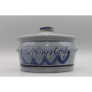 May include: A light blue ceramic crock with a matching lid, featuring a speckled finish and the dark blue hand-painted text Heringstopf on the front. The vessel has two small side handles and a dark blue horizontal band near the base.