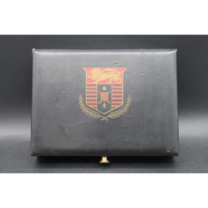 May include: A rectangular black storage box with a hinged lid and a small gold-toned knob at the bottom center. The top of the box features a printed heraldic crest in red and gold, depicting a lion above a shield with a sword and fleur-de-lis.