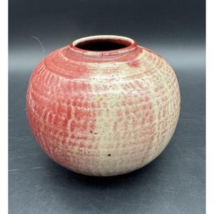 Puede incluir: Un jarrón de cerámica redondo con una abertura ancha y un cuello corto. El exterior presenta una superficie texturizada con un patrón moteado de esmalte rojo intenso y blanco crema, mostrando variaciones sutiles y pequeñas manchas.