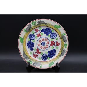 May include: A round ceramic plate featuring a central pattern of red floral motifs surrounded by blue leaves and flowers. The outer rim displays a yellow border decorated with illustrations of grey rabbits and green foliage on a white background.
