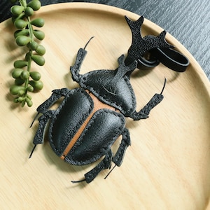 May include: A black leather beetle keychain with a textured surface and a tan center stripe. The insect-shaped charm features a prominent horn, articulated legs, and a black leather loop attachment at the top for securing to keys or bags.