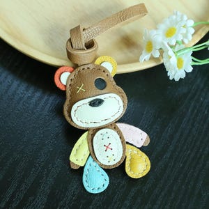 May include: A brown leather bear keychain featuring a patchwork design with multicolored limbs in yellow, light blue, and pink. The bear has a white muzzle, a black eye patch, a yellow stitched X on one ear, and a central belly patch with colorful stitching.